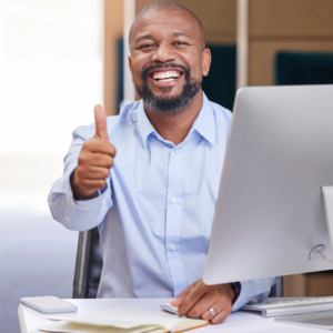 A smiling man giving a thumbs up gesture while sitting at a desk with an open laptop.